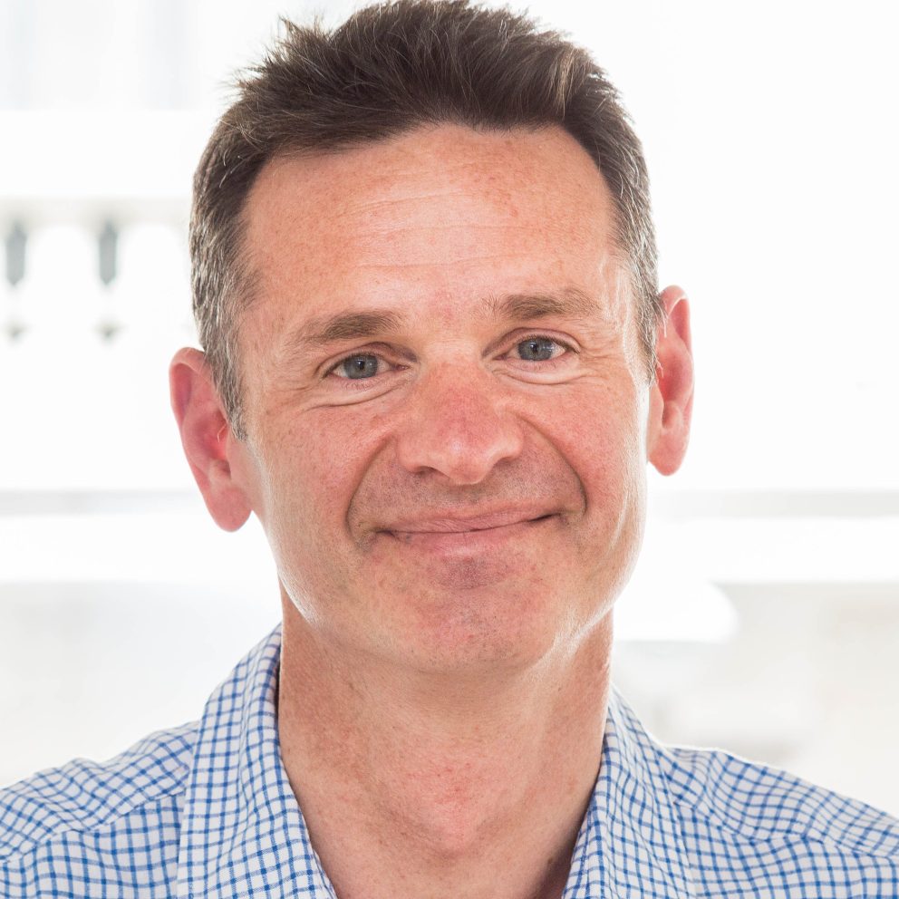 John Drew Profile Pic Smiling In Open Necked Blue And White Checked Shirt With Light Background