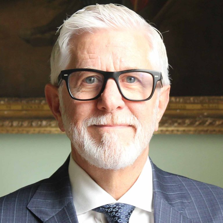 Michael Cook portrait in jacket and tie with framed picture behind