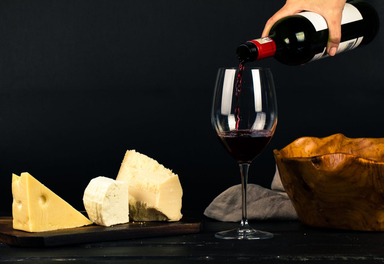 A woman with pink nail varnish pours a bottle of deep red wine into a tall wine glass that sits on a table surrounded by different cheeses