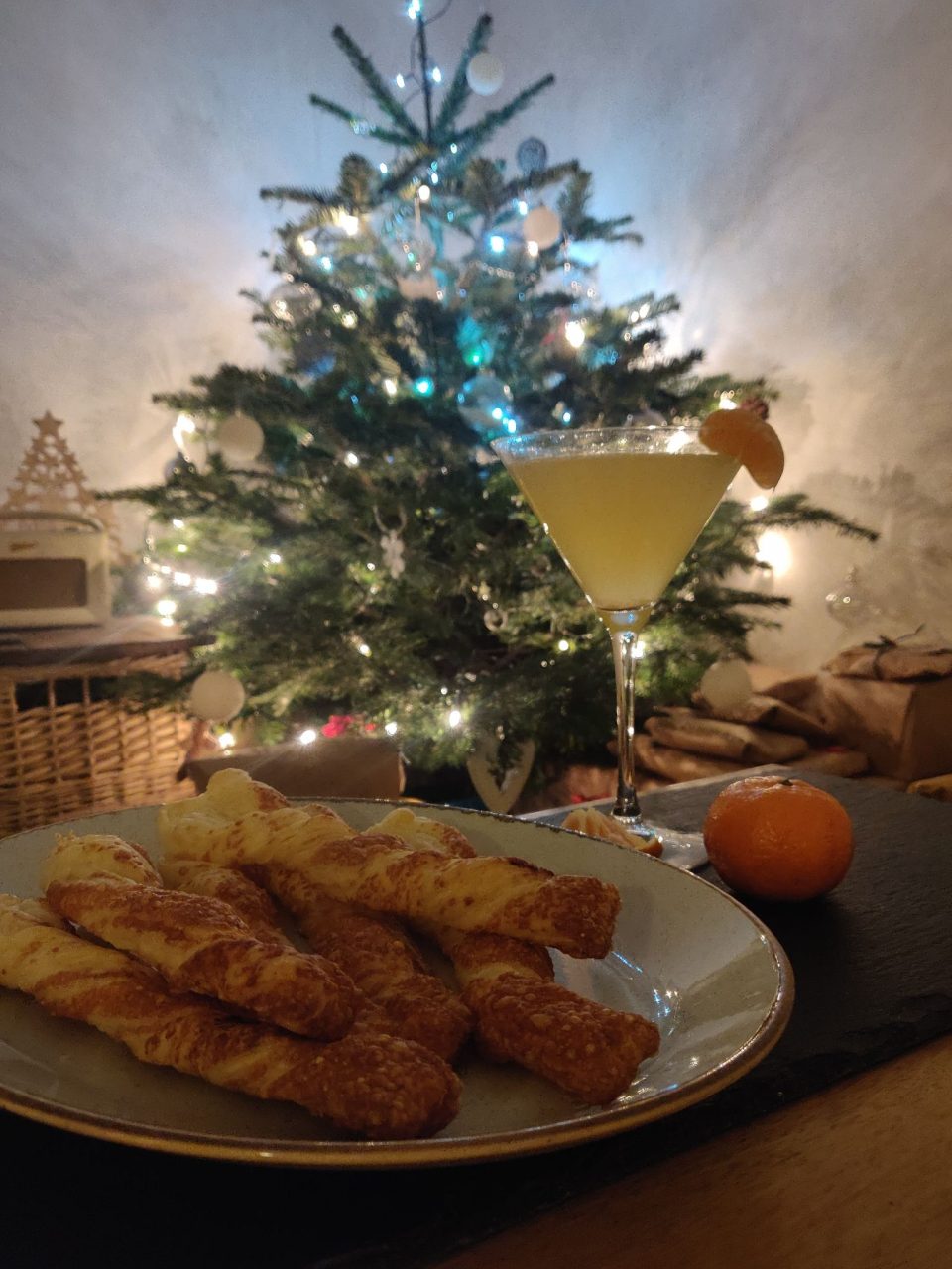 A martini glass of orange liquid and a plate of freshly baked cheese straws in front of a sparkling Christmas tree