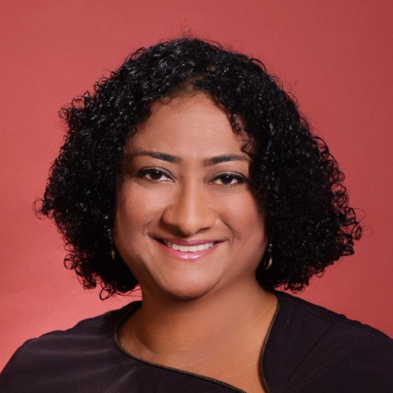 Meera Selva headshot smiling to camera with dark top and red background