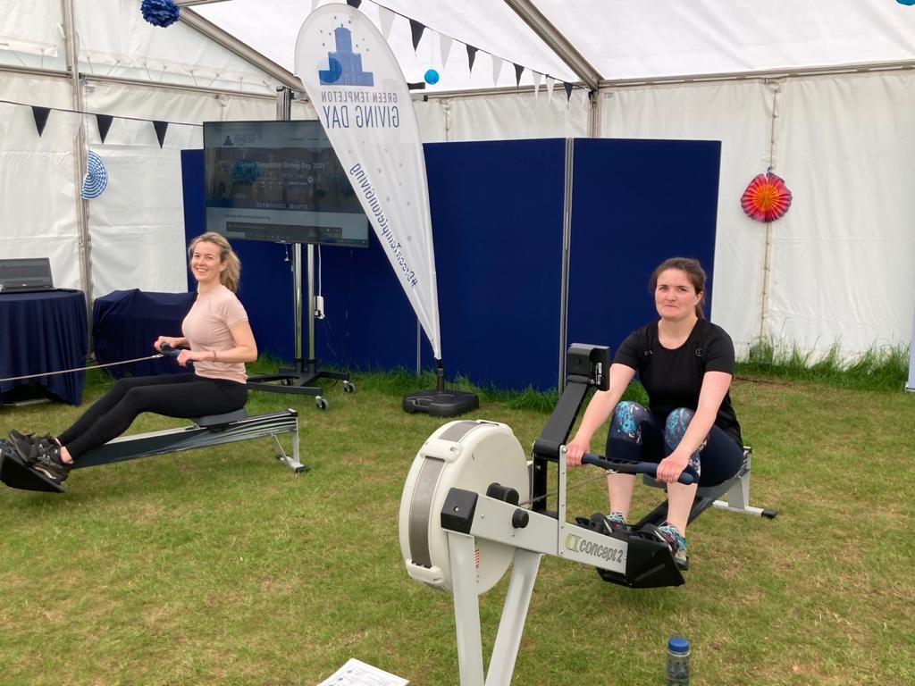 Students Danielle Henriquez and Jacqueline Murphy row side-by-side on machines in the garden marquee