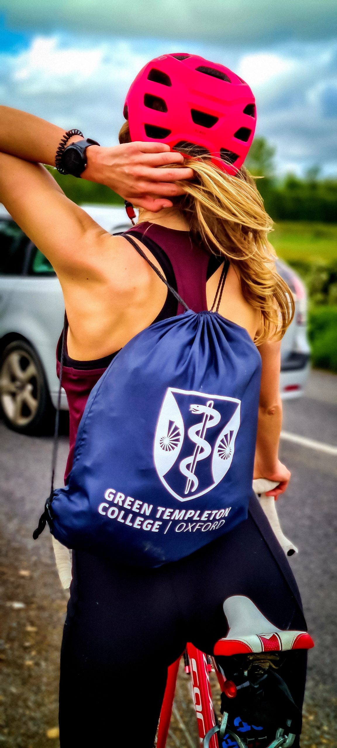 A cyclist in a tank top and pink helmet wears a GTC branded backpack as she takes a break on a long journey
