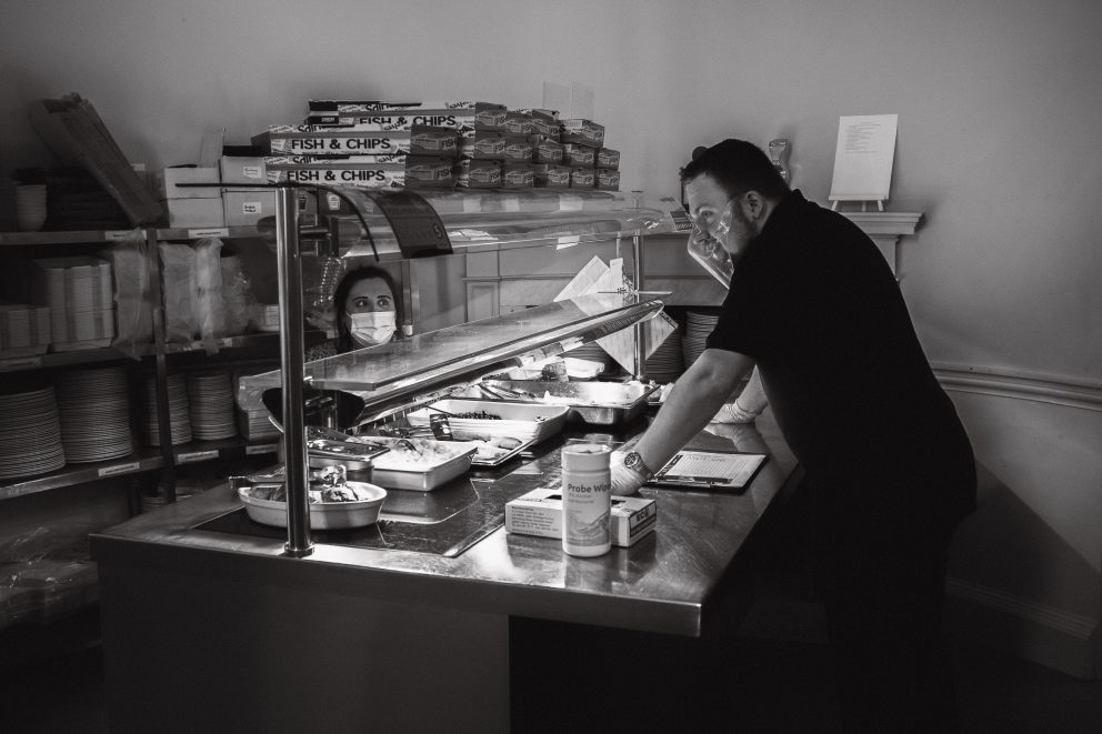 Hannah and Anton from the catering team are separated by a glass screen as they stand in the servery preparing food.