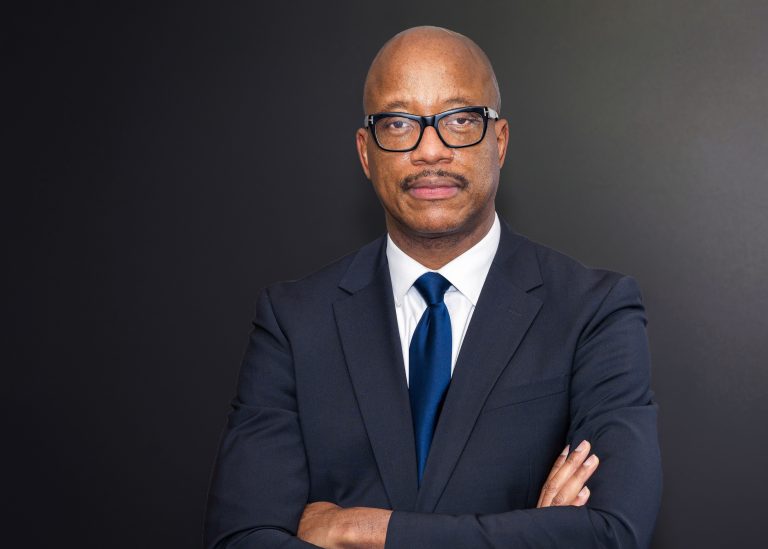Kevin Fenton stood infront of a dark background wearing a navy suit, arms folded, wearing glasses.n2022