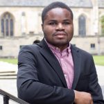 Ebenezer Azamati, leaning on a railing they wear a suit jacket and red checked shirt.