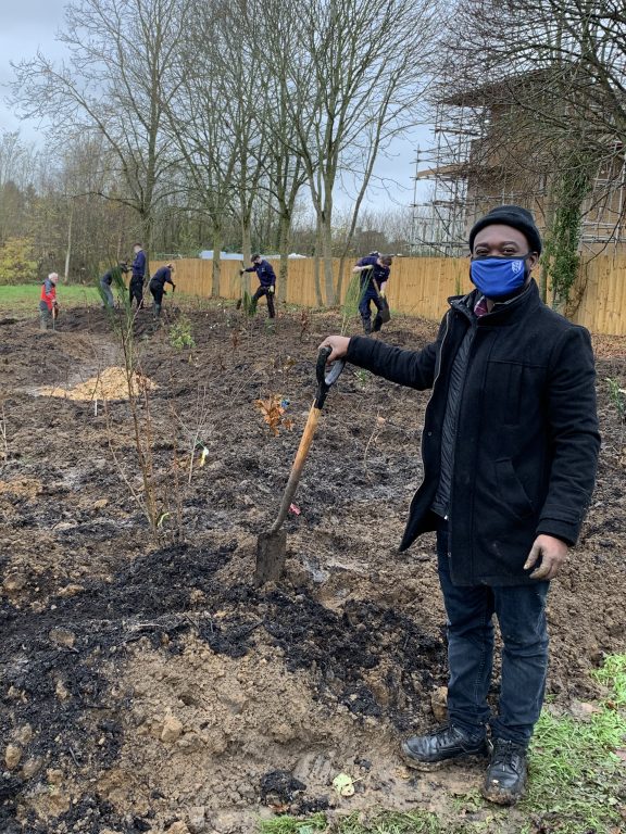 Acheampong Atta-Boateng in coat, hat and face covering holding spade with others behind also on site of planting