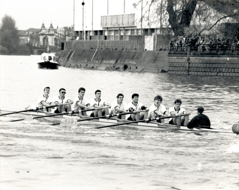 Cal Maclennan rowing in eight-seat boat with cox in middle of river with boat behind in black and white