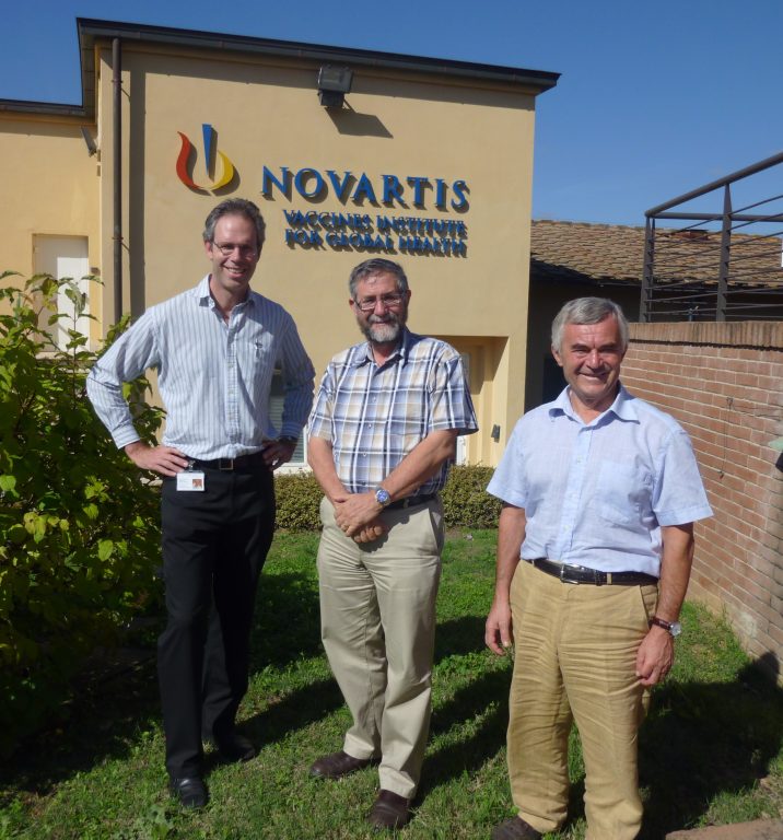 Cal Maclennan on grassy area in front of building with NOVARTIS logo alongside two collaegues all in open-neck shirts on sunny day