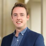 Josh Brewin headshot pictured inside wearing open collar blue shirt and dark jacket
