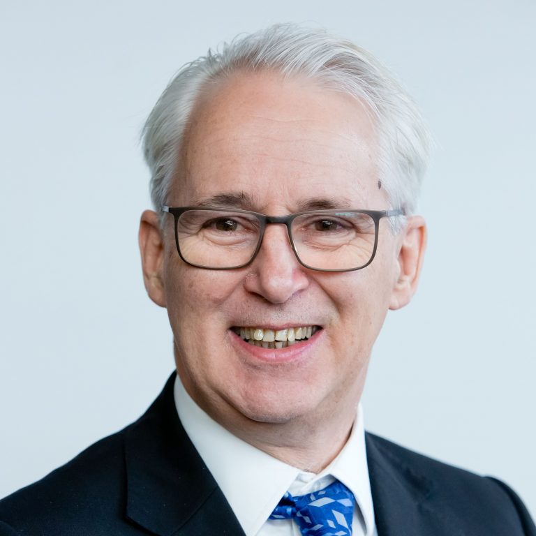 Simon De Lusignan smiling headshot wearing dark suit, white shirt and bright blue tie cropped square