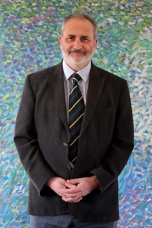 Laurence Leaver In dark jacket and tie and blue trousers in front of abstract painting in green and blue with bits of red