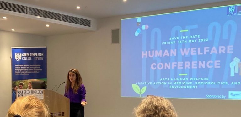 Co Chair Giada Welcoming Attendees to the Human Welfare Conference 2022 from behind lectern with branded popup banner and projected slide behind