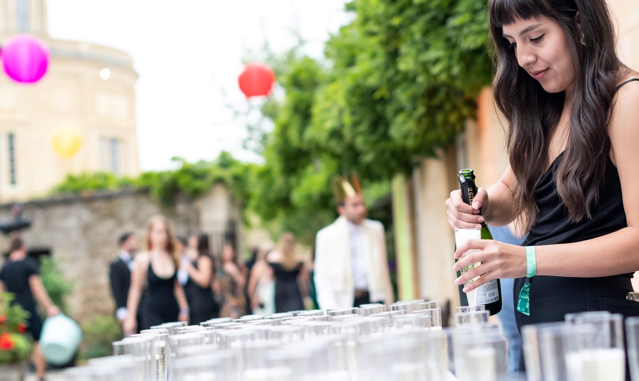 Individual Pouring A Glass Of Fizz Above Table With Lots Of Glasses With Smartly Dressed Ball Guests Behind And Radcliffe Observatory In Background
