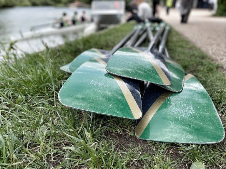Blades of oars gathered in foreground on grass by river with boat with rowers seated in background
