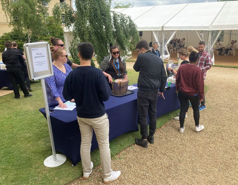 Catering staff serving guests at BBQ lunch in gardens with people seated around marquee behind