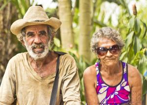 An ageing man in a brown T-shirt and hat sits next to an elderly woman with a purple vest and sunglasses