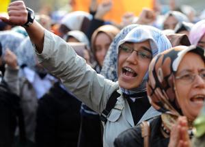 A woman in glasses and a head scarf raises her arm in the air as she walks with other women