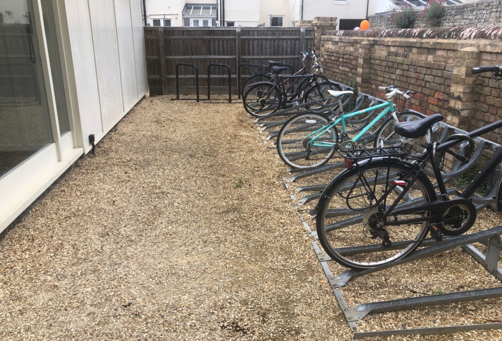 Four bikes on large rack on gravel with building to left and brick wall to right