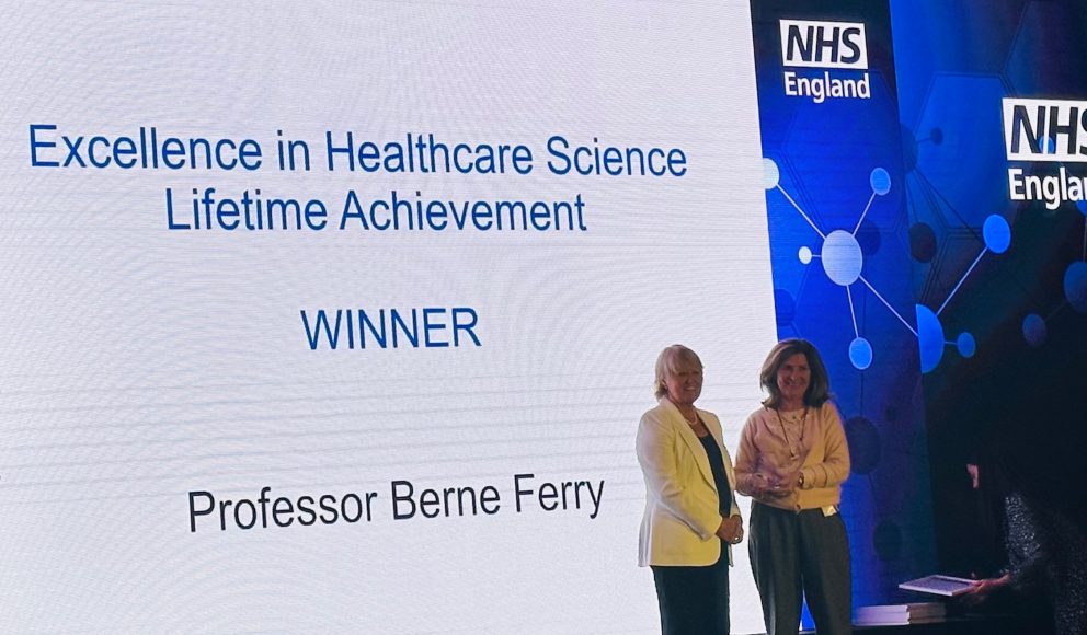 Chief Scientific Officer For NHS England Sue Hill And Berne Ferry Accepting Award on stage with screen and NHS logo behind