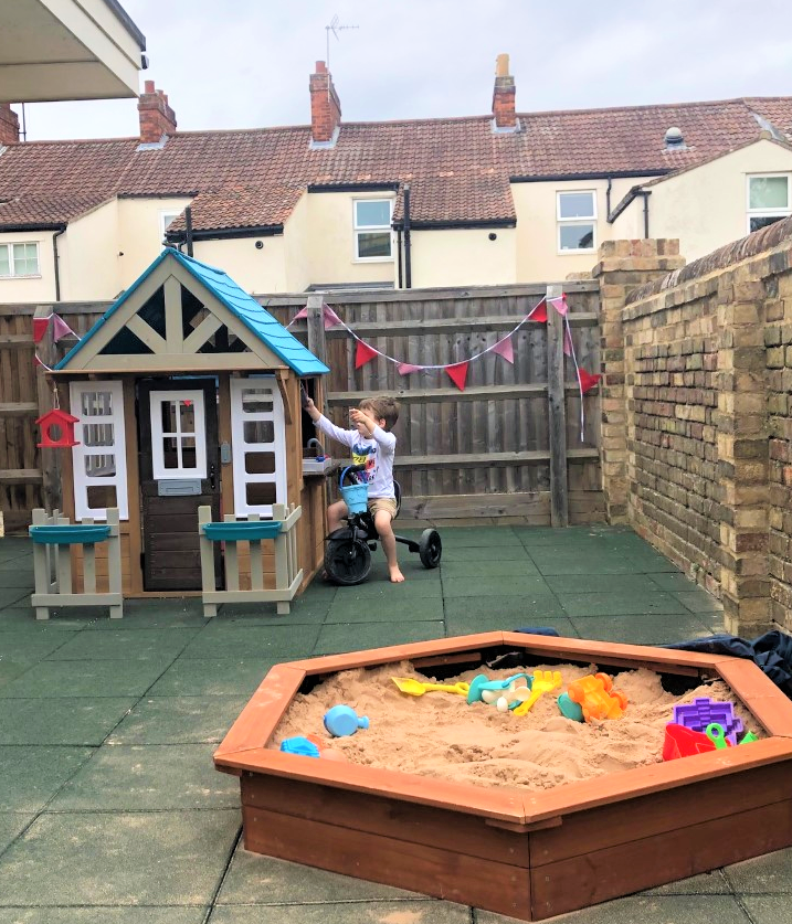 Child on trike playing near little house with sandbox in foreground