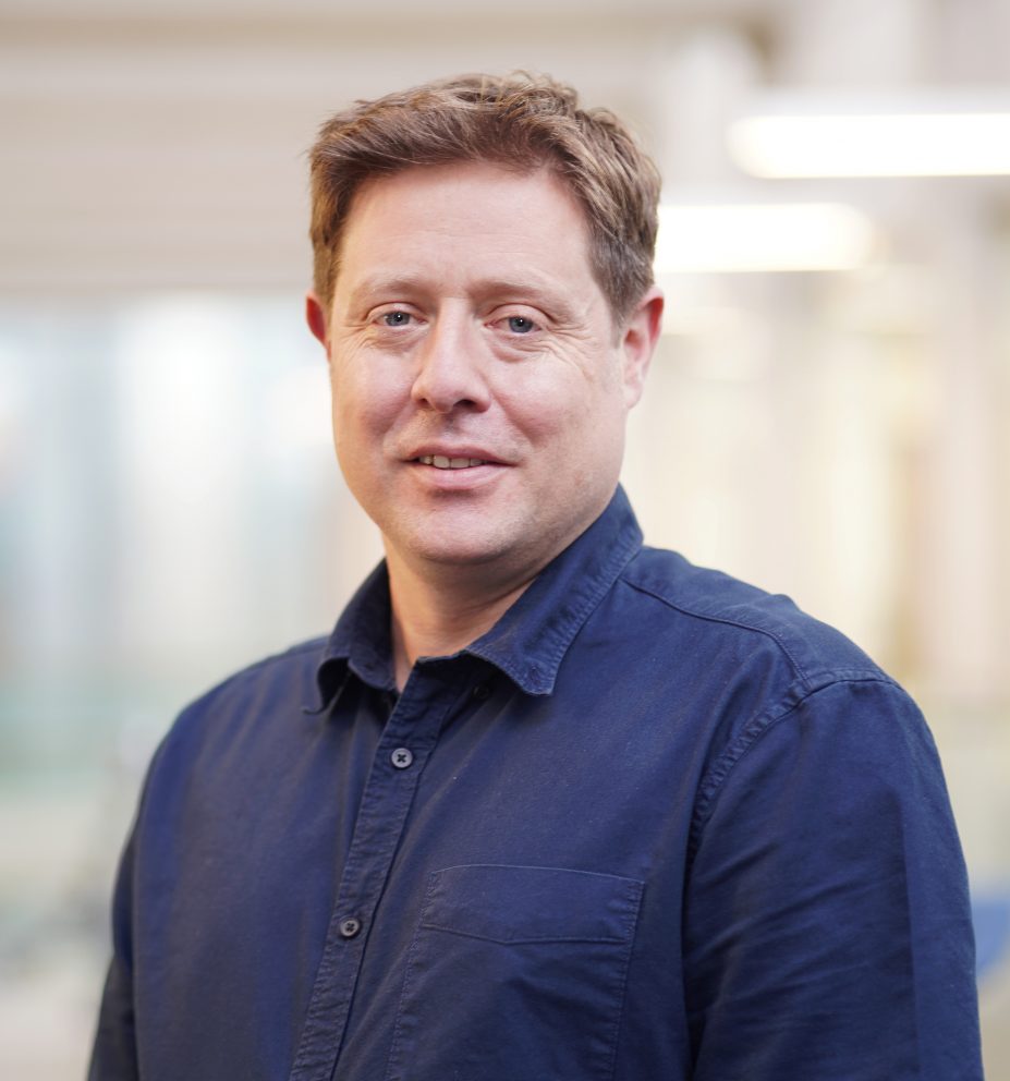 Jon Glasby Headshot in dark blue open-neck shirt