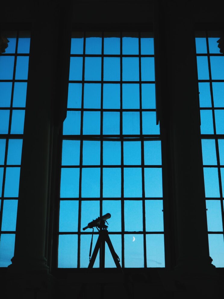 Large high window frame with blue light fading to near darkness with moon framed in one pane and telescope centred at bottom of window
