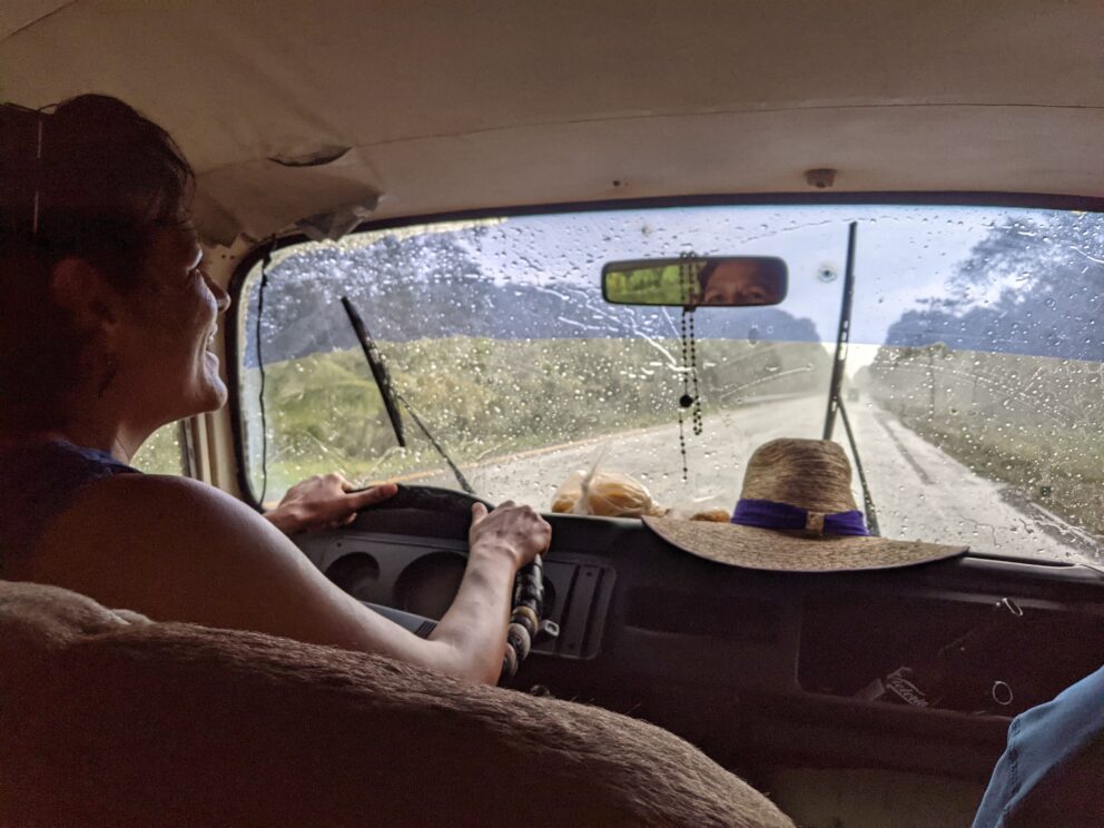 Individual driving in car with windscreen wipers clearing rain and open road ahead. Hat on dashboard in front of driver.