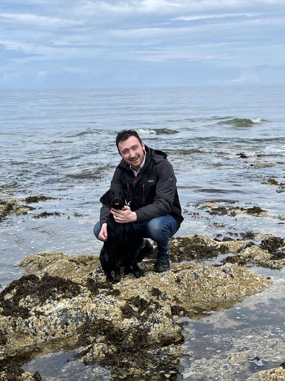 Stefan Tucker knelt down on a rocky coastal terrain with a black Labrador 