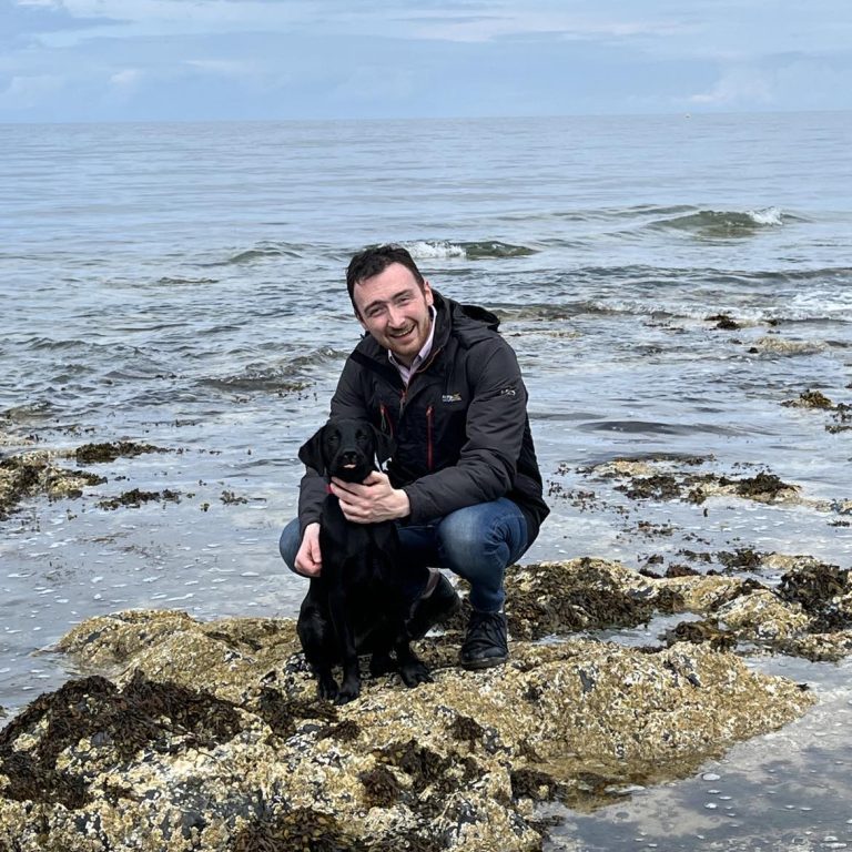 Stefan Tucker knelt down on a rocky coastal terrain with a black Labrador