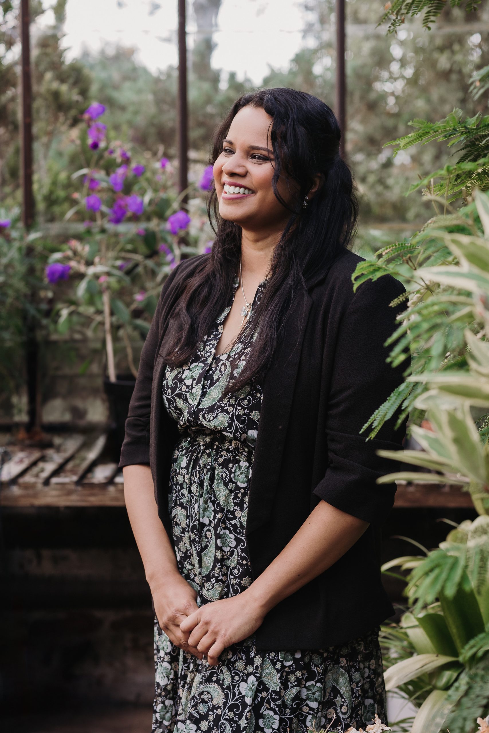 Dr Shobhana Nagraj stood in greenhouse looking off camera surrounded by greenery and flowers