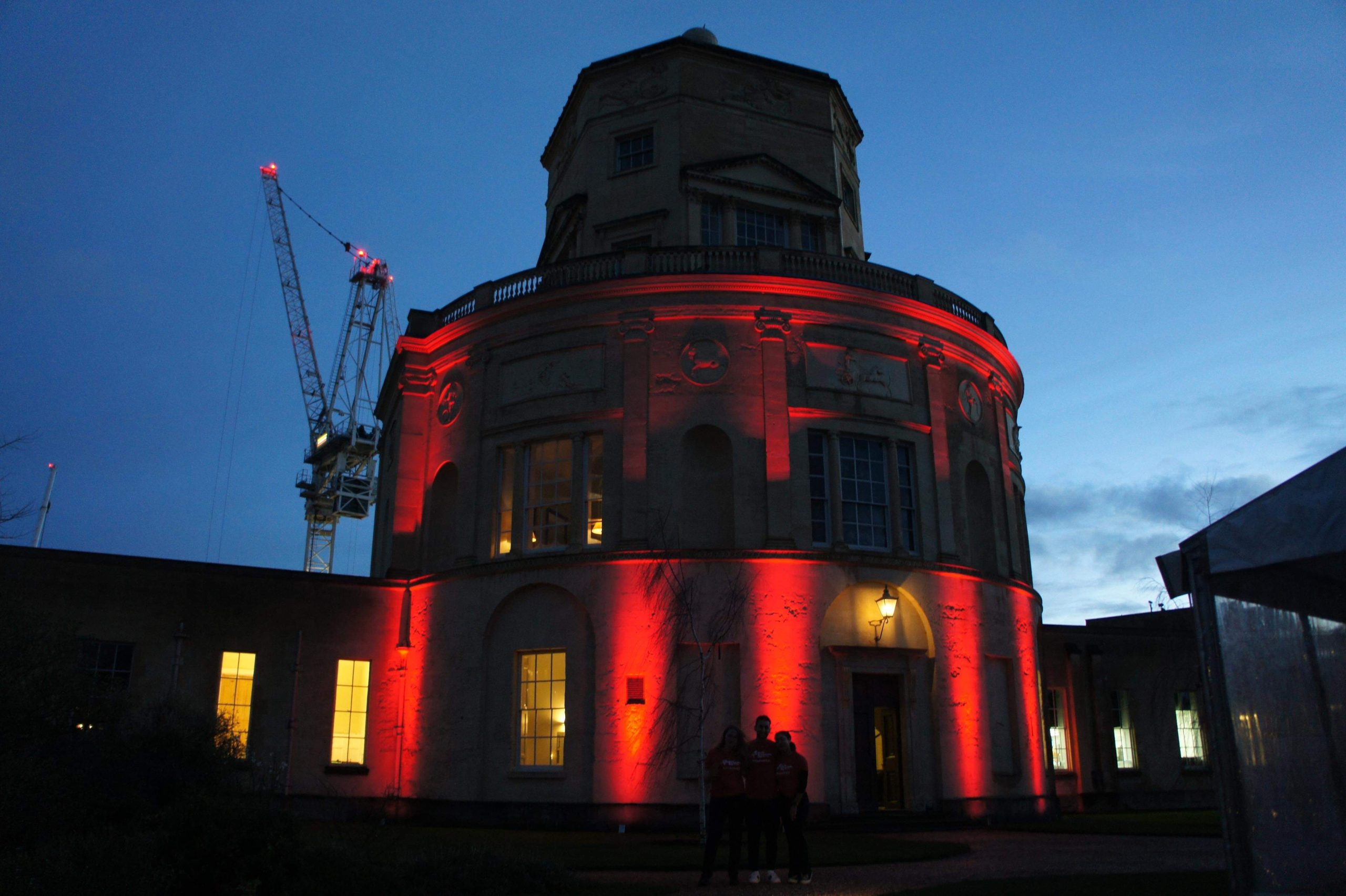 Radcliffe Observatory Lit Red With Crane Behind And Three Individualsin Front In Red T Shirts