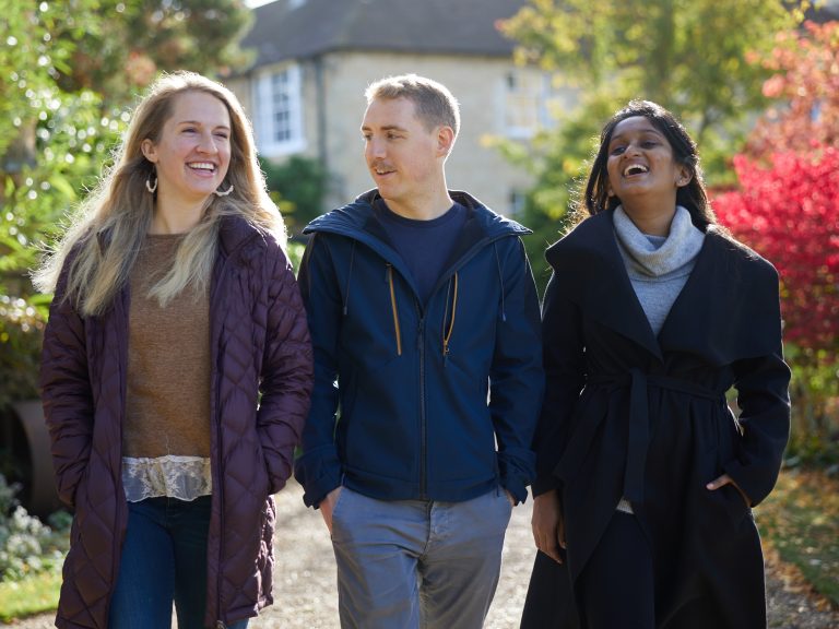 Three Students Walking Through Gardens On Bright Autumnal Day In Coats