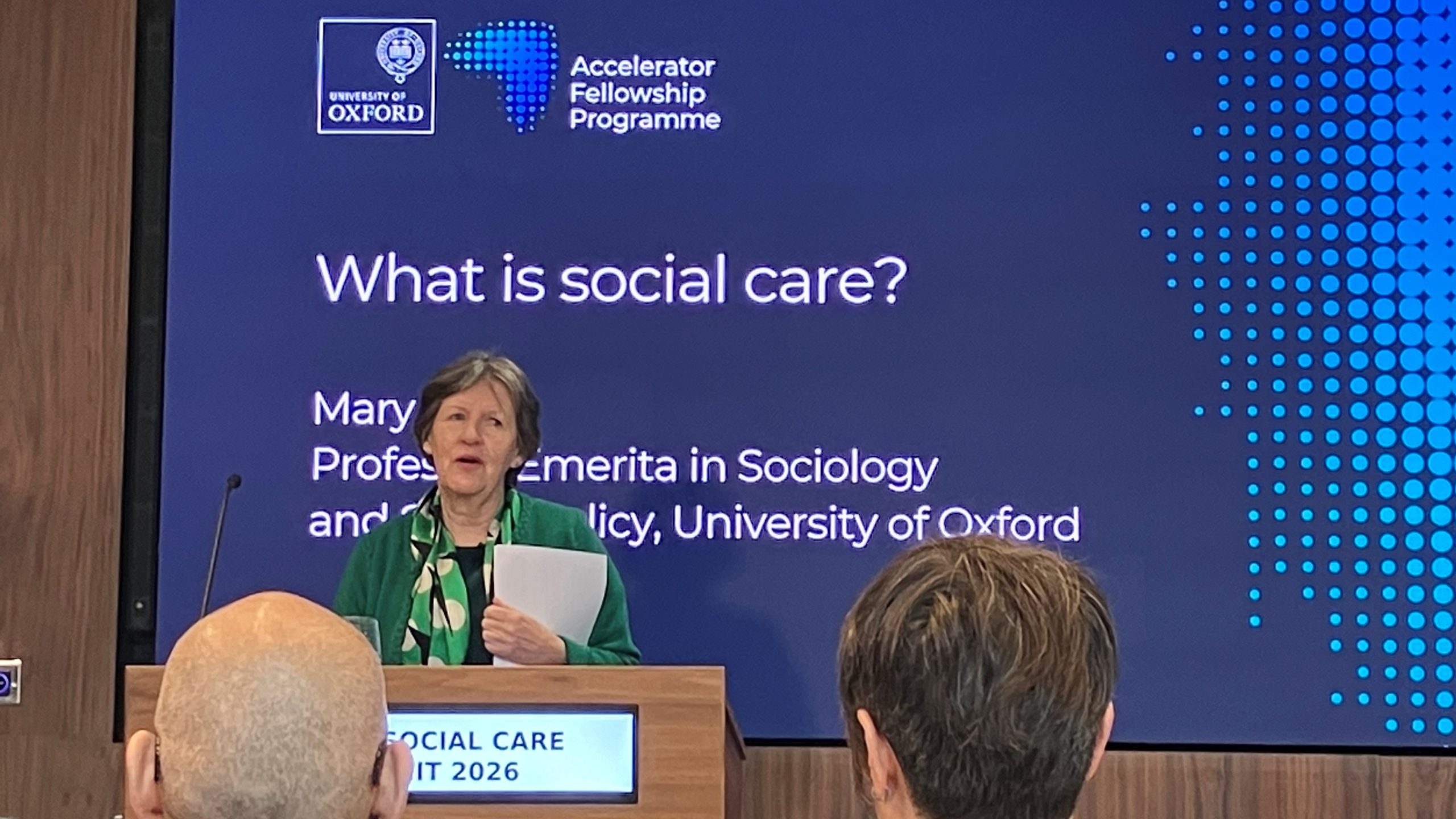 Mary Daly Speaking At Lectern With The Title What Is Social Care Projected On Screen Behind
