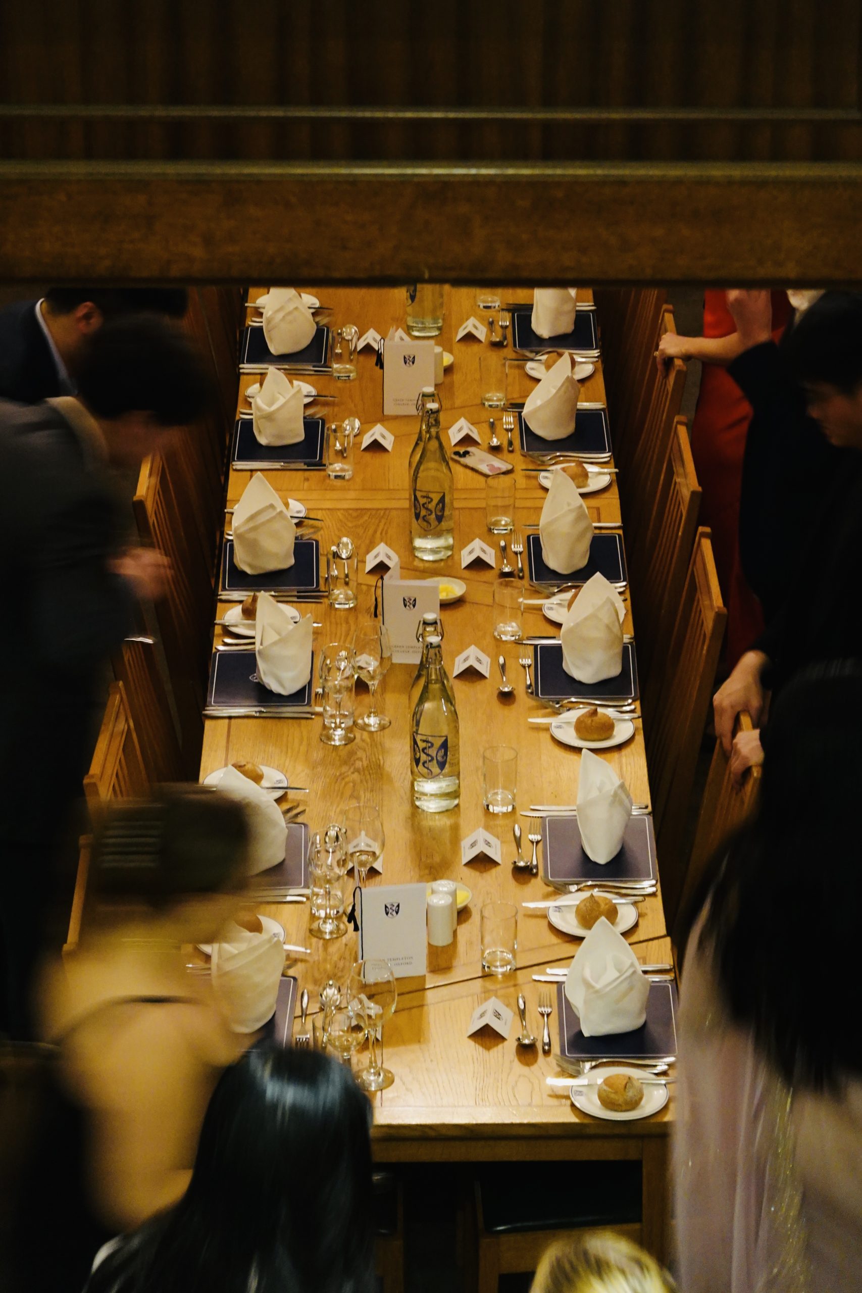 A long formal dining table set for a banquet is viewed from above through a gap in dark wooden panelling. White folded napkins, place settings, water bottles and bread rolls are neatly arranged, while blurred guests move into their seats around the table, creating a sense of activity before the meal begins.