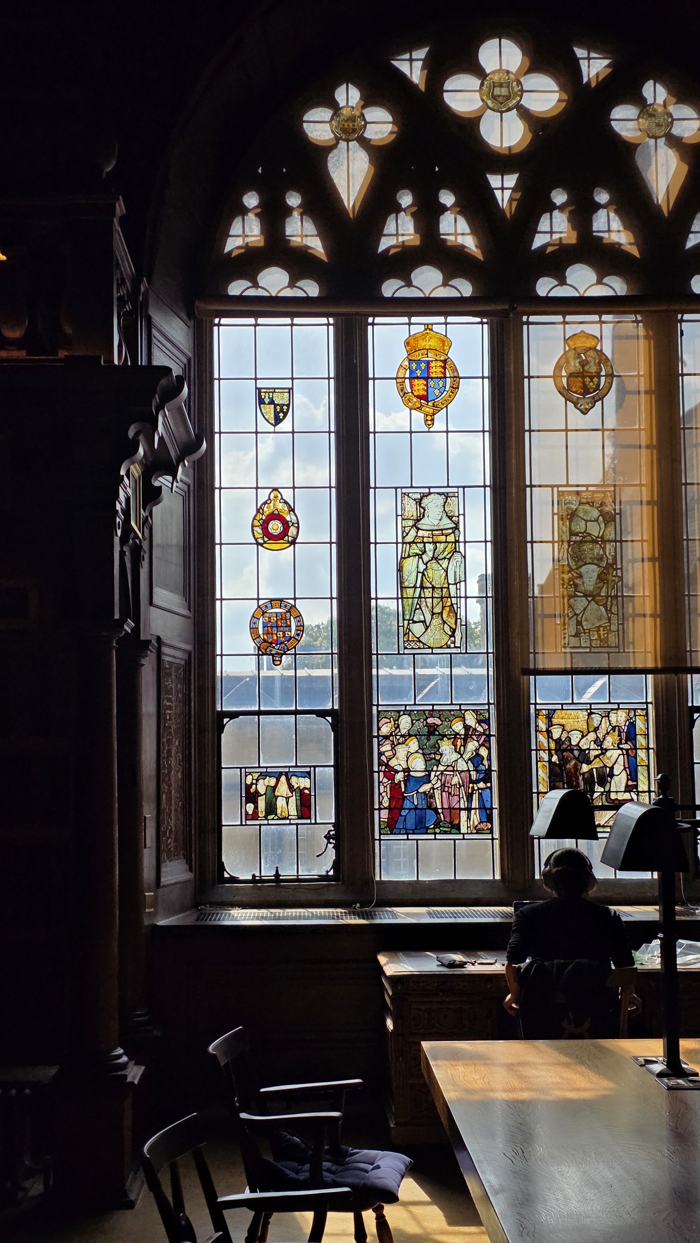A person sits at a wooden desk in a historic library or reading room, silhouetted against a large stained-glass window. Sunlight streams through the decorative glass panels, which feature heraldic emblems and illustrated scenes, illuminating the dark wood interior and study tables.