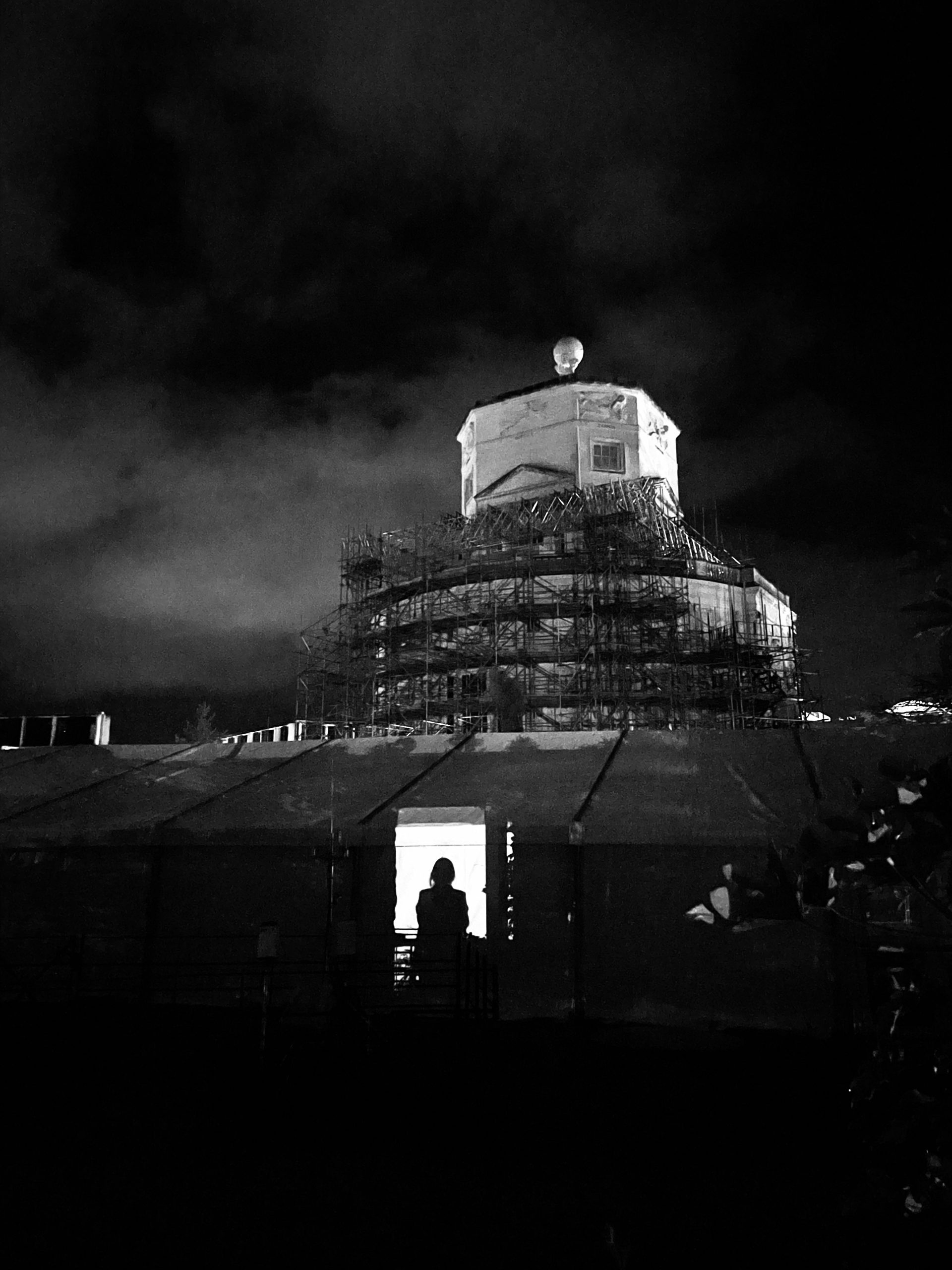A black-and-white night photograph of a domed historic building surrounded by scaffolding during restoration work. Low clouds drift across the dark sky above, while a silhouetted figure stands in a brightly lit doorway of a temporary structure in the foreground.
