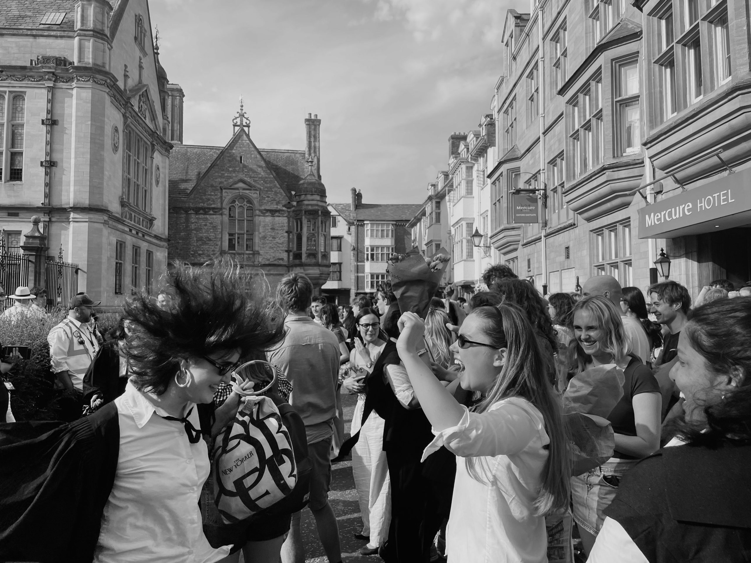 A crowd of people gathers on a historic city street lined with stone buildings. In the foreground, a person in academic dress laughs as their hair is blown dramatically into the air, while friends and onlookers smile and celebrate around them.