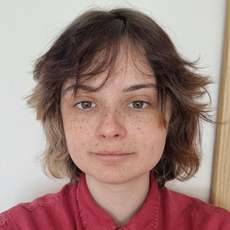A person with short brown hair, freckles and a red collared shirt looking at the camera against a plain light background.
