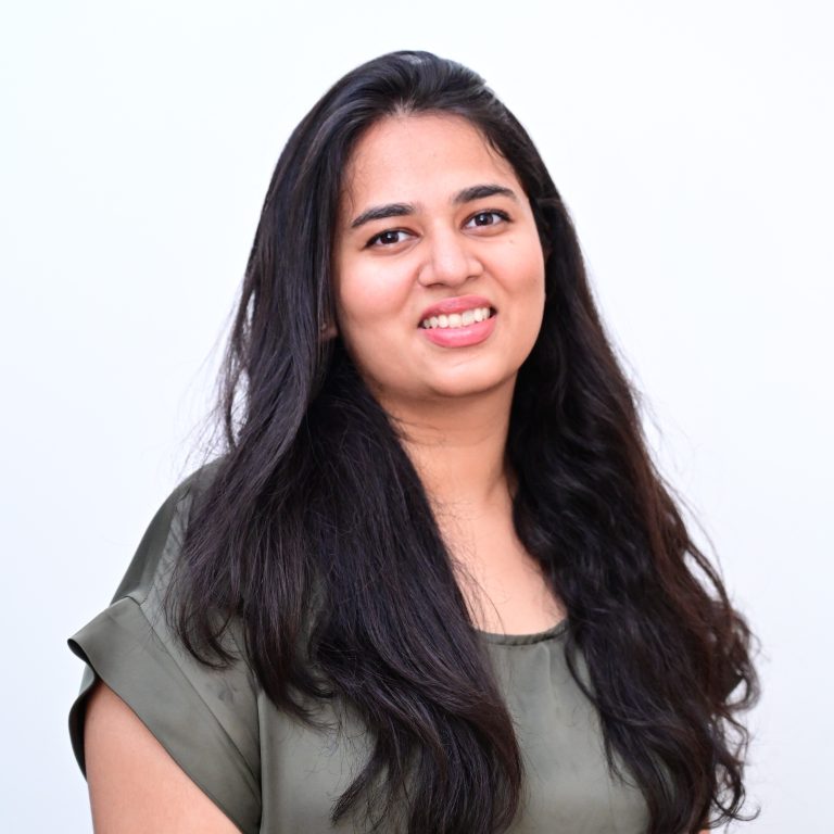 A smiling person with long dark hair wearing an olive green top against a light grey background.