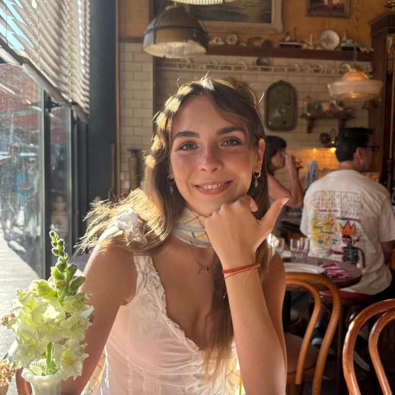 A person in a white top smiles at the camera while seated by a sunlit café window, resting their chin on their hand with a vase of white flowers in the foreground.