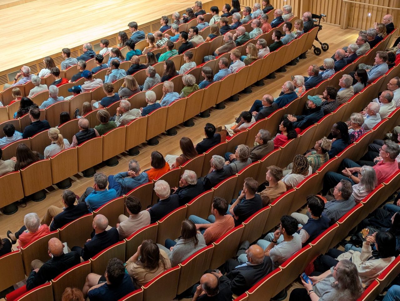Large auditorium with rows full of people seated, looking towards what appears to be a stage on the left